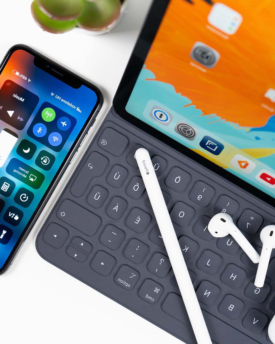 cell phone iPad with keyboard, white stylus, white AirPods; and an iphone on table mobile phone